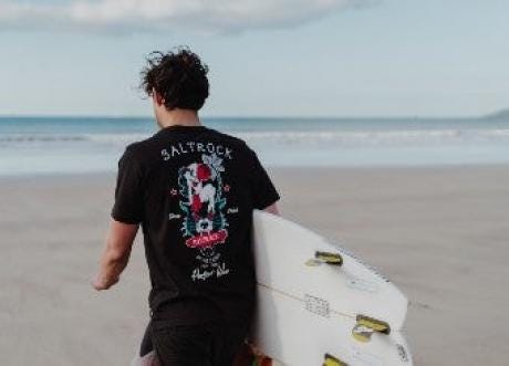 Man in saltrock t shirt walking on beach carrying a surf board